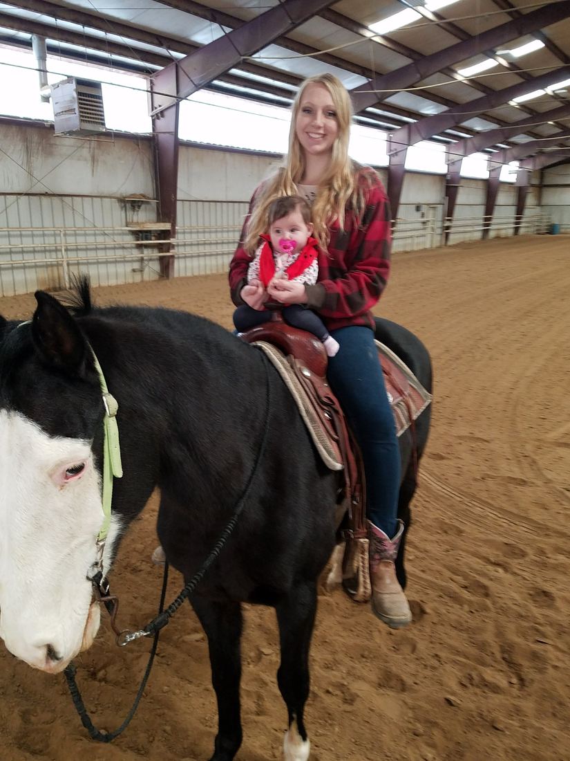 Alena on a black and white horse holding her baby daughter in the saddle, first season